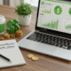 Laptop showing MyGreenBucks dashboard with financial charts, notebook with Kenneth Jones' program name, and office accessories on wooden desk